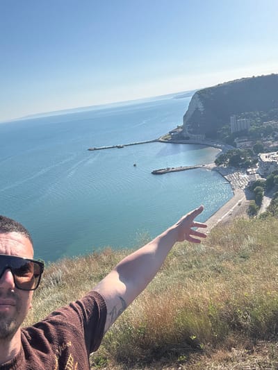 The media file is a selfie taken outdoors, with the photographer's arm extended and hand open. The scene is dominated by a vast expanse of calm, blue water, likely the sea, which reflects the bright sunlight. In the background, a rugged cliff face rises, dotted with buildings and lush green vegetation. The top of the cliff is obscured by a hazy sky, suggesting a clear day with some atmospheric diffusion.
The photographer is wearing sunglasses and a brown, textured shirt. A tattoo is visible on their arm. The overall impression is one of a person enjoying a scenic view, possibly on vacation or during a hike. The activity taking place is the act of taking a photograph.
Notable details include the clear, bright daylight, indicating it is likely mid-morning or afternoon. The location appears to be a coastal area with significant elevation, offering a panoramic view of the water and cliffs. The open hand gesture could be interpreted as a sign of greeting, excitement, or simply to frame the view.
There is no visible text within the image. The setting suggests a natural landscape combined with a developed area, possibly a town or resort situated on the coast.