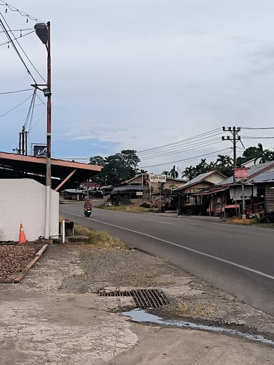 This image captures a roadside scene in Sukamulia, Indonesia, under an overcast sky. A two-lane paved road, marked with a single white line, extends into the distance. On the left, a cracked concrete shoulder features a metal drain grate with a small puddle, and an orange traffic cone. Behind this, a low white wall fronts a building with an exposed rafter roof, and a large utility pole heavily laden with wires and streetlights.

A person on a red motorcycle, wearing a head covering, is seen riding away from the viewer along the road. On the right side of the road, a series of modest, single-story buildings, constructed from wood and possibly concrete, line the street. These structures appear to be homes or small businesses. Another utility pole with multiple power lines stands further down on the right.

Lush green trees, including palm trees, are visible behind the buildings and in the background. Signs with visible text include "LA" on a black banner to the left, and further down the road on the right, "GADING GAJAH" and "MEUTUAH" are displayed on storefront signs. The overall atmosphere suggests a quiet, everyday moment in a semi-urban or rural setting.