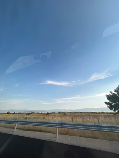 The image depicts a roadside scene with a vast expanse of sky and a distant landscape. The sky is a prominent feature, taking up a significant portion of the frame. Wispy white clouds are scattered across the blue expanse, catching the light. The overall impression of the sky is bright and clear, suggesting fair weather.
In the foreground, the glossy surface of what appears to be a car hood reflects the sky and clouds, creating a mirror-like effect. This reflection is slightly distorted, adding an interesting visual element.
To the right of the sky and reflection, a metallic guardrail runs horizontally across the mid-ground, separating the road from a field. The field beyond is covered in dry, yellowish grass, indicating a rural or agricultural setting. In the far distance, a range of mountains is visible, their outlines softened by the haze. A few trees are scattered along the edge of the field and in the distant landscape.
The road itself is visible on the right edge of the image, with a strip of asphalt and a concrete curb. The presence of the guardrail and road suggests this is a highway or a similar type of road.
The lighting indicates it is likely daytime, possibly late afternoon or early morning, given the soft quality of the light and the angle of the sun. There are no people or specific activities clearly depicted, making it a static scene. The location cues, such as the guardrail and the type of landscape, are consistent with the provided location context of Saraia, Bulgaria. No text is visible in the image.
