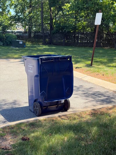 This image captures an outdoor scene, likely a suburban residential area, dominated by a large, dark blue trash or recycling bin. The bin is positioned on a light grey paved surface, such as a driveway or street, next to a concrete curb that separates it from a vibrant green grassy strip.
The background features lush green trees with dense foliage, suggesting a summer or spring setting. Behind the trees and grass, a dark wooden fence is visible. To the left side of the frame, a brown, metal signpost stands, holding a small, rectangular white sign. The text on this white sign is unreadable due to blurriness. Further back and to the right, nestled among the trees and near the fence, is a green utility box.
The lighting indicates a bright, sunny day, likely late morning or early afternoon, as distinct shadows are cast by the bin and trees onto the paved ground. The overall weather appears clear and pleasant.
There is no discernible activity or event taking place; the scene appears static.
Visible text provides location information: on the side of the blue bin, the words "PROPERTY OF", "TOWNSHIP", "OF", and "LAWRENCE" are clearly legible, identifying the bin as belonging to the Township of Lawrence. On the green utility box, the number "8674" is visible. The scene strongly suggests a typical street or residential property within Lawrence Township, United States of America.