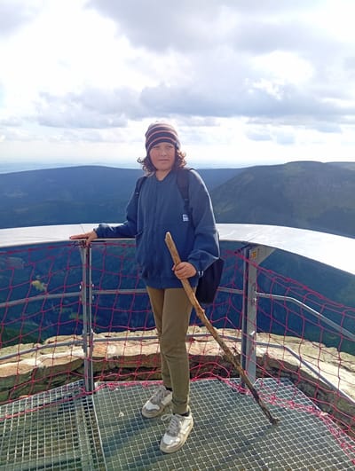 A person stands on an observation platform overlooking a mountainous landscape. The person is wearing a blue hooded jacket, khaki pants, a striped beanie, and white sneakers. They are holding a wooden stick. The observation platform has a metal grate floor and a railing with a red safety net. In the background, rolling hills and mountains stretch to the horizon under a cloudy sky. The scene appears to be the Sněžka mountain peak in Malá Úpa, Czechia, a popular tourist destination known for its panoramic views.