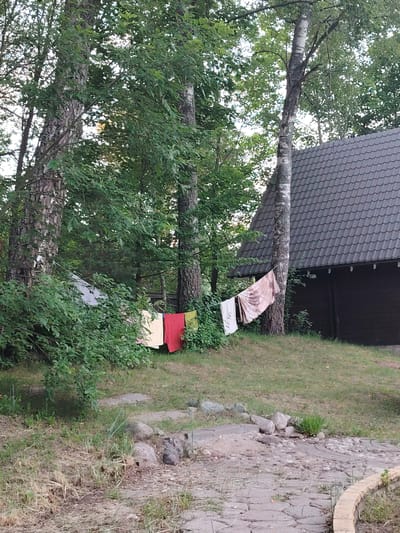This media file depicts an outdoor scene near a house with a dark, shingled roof, nestled among trees. A clothesline stretches across the foreground, adorned with several colorful pieces of laundry, including a red towel, a yellow garment, and a patterned fabric. The laundry appears to be drying in the sun.

The setting is a grassy area with a stone path leading into the frame. Several large rocks are scattered on the ground, and dense green foliage surrounds the scene, suggesting a natural or semi-rural environment. The trees are tall and leafy, with one prominent birch tree with distinctive white bark standing near the house.

The lighting suggests it is daytime, likely late afternoon or early evening, as the light is soft and diffused. There are no people visible in the frame. The overall mood of the image is peaceful and domestic, evoking a sense of everyday life in a quiet setting.

The location context provided is Мётчанскі сельскі Савет, Belarus, which places the scene within a specific administrative region of Belarus. There is no visible text in the image.