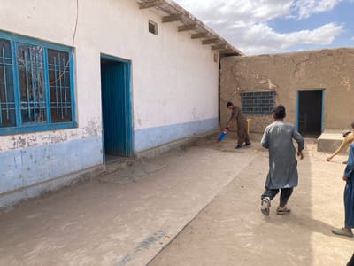 The image shows a group of young men playing cricket in a dirt courtyard in Saranan, Pakistan. The courtyard is located between two buildings, both of which are made of brick. The building on the left has a large blue window and a blue door, while the building on the right has a smaller blue window and a blue door. The courtyard is dusty and there is a yellow plastic bucket in the corner. One of the men is swinging a red and blue cricket bat while another man is running towards the bat. A third man is watching the game, while a fourth man is walking in the background. The sky is blue with white clouds. The image was taken at 30.532° N, 66.909° E.
