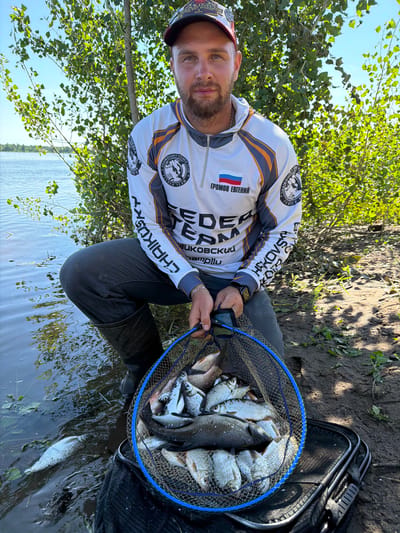 A Caucasian man is crouched by the water's edge, holding a fishing net full of fish. He is wearing a long-sleeved fishing shirt with orange and white accents, dark pants, and black waders. The shirt has graphics including what appears to be a Russian flag and Cyrillic text. He has short brown hair and a beard. The fishing net is blue and contains numerous silver-colored fish of various sizes. A black fishing bag is visible next to the net. The man is positioned on a muddy bank beside a body of water, with trees and foliage in the background under a clear blue sky. The scene appears to be a natural outdoor setting, likely a riverbank or lake shore, in or near Kambarka, Russia.