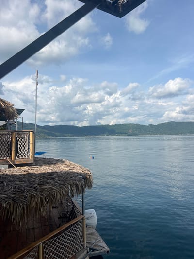 The image depicts a daytime view from a waterside structure in Unknown, El Salvador, overlooking a large body of water with distant hills. In the foreground, the lower left and bottom portions show a wooden deck with a woven rope railing and a thatched roof made of dried fronds. A white "SUZUKI" outboard motor is mounted on a wooden dock extending into the water on the lower right. Overhead, a dark metal beam partially obscures the sky in the upper left. A tall flagpole stands vertically to the left of the image center, displaying a furled flag with red, white, and blue segments, consistent with the flag of El Salvador. To the right of the flagpole, a blue object is partially submerged in the water. Further out, a small red and blue floating object is visible in the water. The calm, blue-grey water fills the midground, showing subtle ripples on its surface. The background features a continuous range of lush green, tree-covered hills or mountains stretching across the horizon. Small structures are faintly discernible along the distant shoreline at the base of these hills. The sky above is light blue with scattered white cumulus clouds of varying sizes. No individuals are clearly visible in the scene.