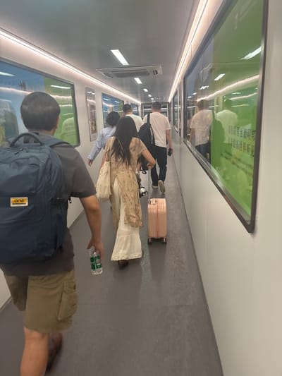 A group of individuals are walking down a brightly lit corridor with large green-tinted windows on the right side. The corridor has a grey floor and white walls, with linear lighting fixtures on the ceiling. The people are walking away from the camera, suggesting they are moving through an airport terminal, train station, or similar transit hub.
In the foreground, a man wearing a dark grey t-shirt, khaki cargo shorts, and a dark blue backpack is walking, holding a clear plastic bottle of water. To his left and slightly ahead, a woman in a floral print dress and a cream-colored skirt is walking, pulling a pink suitcase. Behind them, two other individuals are visible, one wearing a light blue shirt and the other in a white t-shirt and dark pants. Reflections in the windows suggest the presence of other people and structures outside. The overall scene depicts movement and transit within an enclosed, modern environment. The city of Xinwu District, China is provided as the location.