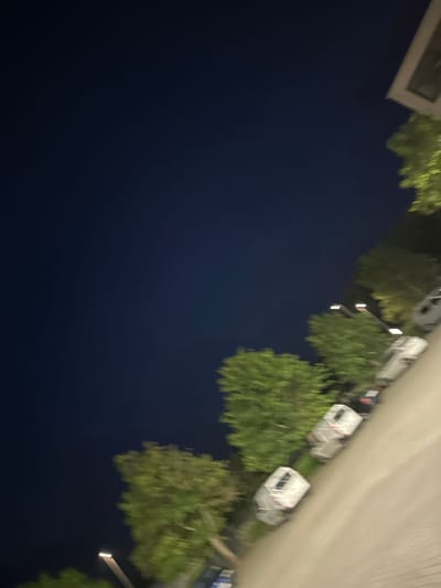 The image depicts a street scene at night, likely in Schweitenkirchen, Germany, based on the provided location. The sky is a deep navy blue, suggesting a late evening or night setting. To the right of the frame, a row of trees with green foliage is visible, illuminated by artificial light sources. Adjacent to the trees, several cars are parked along the side of a paved road. The cars appear to be a mix of white and darker colors, and some are partially obscured by the trees. Streetlights with glowing white tops are also visible, illuminating parts of the scene. The perspective is slightly angled, giving a diagonal view of the street and parked vehicles. The image is somewhat blurry, indicating motion or low light conditions. The overall environment suggests a residential or commercial area where parking is available along a street.