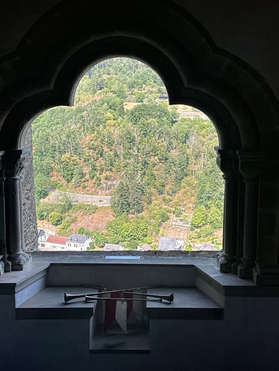 This image, captured from a window in Vianden Castle in Vianden, Luxembourg, presents a view of the town and surrounding hills through an arched, Gothic-style window frame. The window opening is framed by dark stone arches and columns, suggesting the historical architecture of the castle. Inside the window frame, on a stone ledge, rest two long, slender brass instruments, resembling trumpets or horns, crossed over each other. Partially visible beneath them are red and white fabric banners, possibly ceremonial or decorative.

Beyond the window, a verdant hillside is visible, densely covered with trees. A winding road snakes its way up the slope, with retaining walls made of stone. Nestled at the base of the hill and spread across the foreground is a small town. Several buildings with red and grey roofs are apparent, indicative of traditional European architecture. The overall scene conveys a sense of looking out from a historical vantage point onto a scenic European landscape.