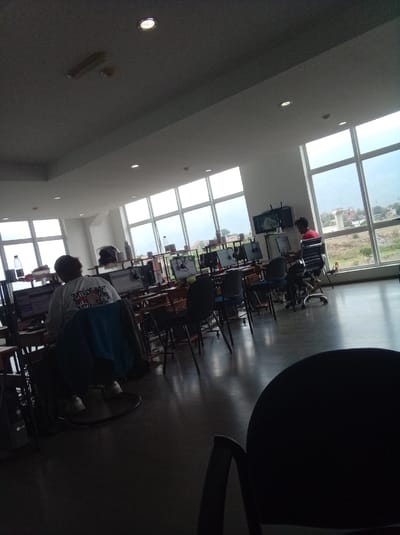 The image shows the interior of a room, likely a library or computer lab, in Parroquia Guatire, Venezuela. The room is filled with rows of desks and chairs, each desk equipped with a computer. People are seated at some of the desks, using the computers. Large windows line the far wall, offering a view of the outside landscape with buildings, trees, and hills. The room has fluorescent ceiling lights and air conditioning vents. A foreground chair is present.