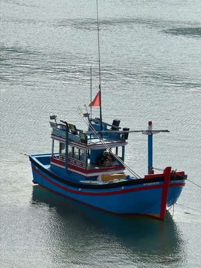 The image, rotated 90 degrees clockwise, displays a vibrant blue and red fishing boat floating on calm, sun-dappled water. The boat is the central subject, positioned with its bow to the right and stern to the left. The surrounding water, reflecting bright sunlight, shows subtle ripples and patterns, indicating clear skies and a serene environment, likely during the day.

The fishing boat features a striking blue hull accented by a prominent red stripe along its upper edge and a narrower black stripe below. The cabin, situated mid-ship, is adorned with decorative red and white panels and visible windows. A mast extends upwards from the deck, displaying a small red flag near its peak. Various ropes, rigging, and what appear to be fishing apparatus or provisions are visible on board.

Visible text on the boat's bow clearly reads "NT 93979 TS", which is likely a registration or identification number and is repeated further along the hull. Partially visible text "UNC-100-Y" is also seen on the side of the cabin structure. The boat casts a distinct shadow on the water, particularly from its forward section. The scene evokes a sense of quietude, with the boat seemingly moored or anchored in a bay or harbor in Ninh Chu Ward, Vietnam.