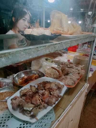 The image depicts a food display inside a glass case, likely a street food vendor's setup in Ea Drăng, Vietnam. A woman is visible behind the top level of the display, which holds a whole cooked chicken or duck and some round objects. The second level of the display case contains several plates and bowls of prepared food. There is a plate of chopped organ meat in the foreground on a white rectangular plate with a lace paper doily underneath it. Behind it, there's a stainless steel bowl with dark-colored meat and a tray of light-colored meat pieces. Stacked bowls are visible on the right side of the second level, along with plastic bags and containers filled with various ingredients. The display case itself appears to be old, with a weathered wooden frame.