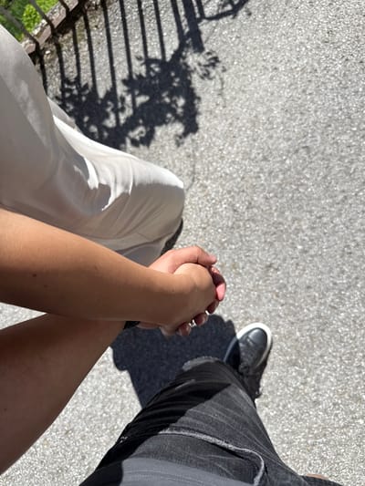 The image shows a close-up, overhead view of two people holding hands while standing on a paved surface. One person, wearing a white dress, has their arm extended and held by the other person, who is wearing dark jeans and black sneakers. The ground is a speckled, light-colored asphalt. In the upper left corner, a dark metal fence casts shadows on the ground, along with the dappled shadows of tree leaves. The scene is outdoors, suggesting a daytime setting with bright sunlight. The image does not provide any explicit visual cues to confirm the location as Shiroka Laka, Bulgaria; the description relies solely on the visual elements present in the photograph.