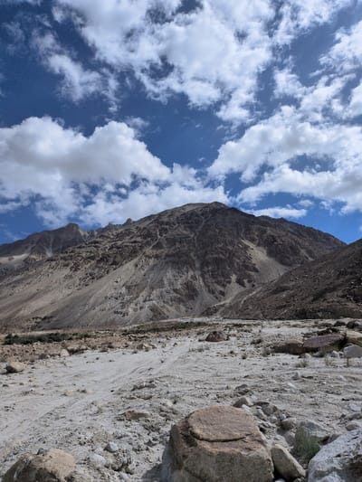 The image depicts a vast, rocky, and sandy terrain with a mountain range dominating the background. The sky above is a vibrant blue, dotted with scattered white clouds. In the foreground, numerous stones and boulders of varying sizes are strewn across the dry, light-colored ground, which shows faint tire tracks. The mountain slopes are rugged and arid, with visible geological layers and shadows suggesting uneven terrain. The scene appears to be a natural landscape, likely in a mountainous region. There are no people or man-made structures visible in the image. The location is identified as Sasoma, India.