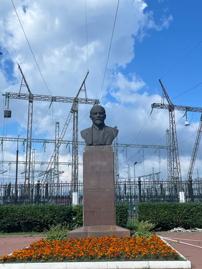 A bust of Vladimir Lenin stands on a granite pedestal in front of a substation in Zavolzhye, Russia. The bust is dark and weathered, with Lenin depicted with a stern expression. The pedestal is rectangular and made of polished granite. Behind the bust, a complex network of electrical towers, wires, and insulators stretches across the frame under a blue sky with scattered clouds. A black metal fence runs along the edge of the substation, separating it from the foreground. The foreground features a bed of vibrant orange marigolds and a neatly trimmed green hedge. The overall scene is one of industrial infrastructure juxtaposed with a historical monument and natural elements.