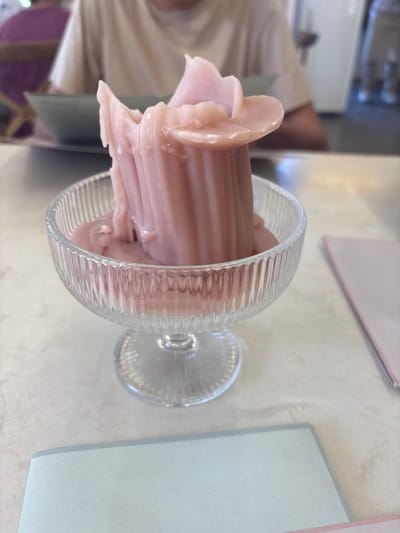 A close-up, slightly high-angle shot shows a clear glass dessert dish filled with a pale pink soft-serve ice cream or frozen yogurt. The glass has a short stem and a fluted design around the bowl. The ice cream is swirled in a way that creates a column of soft-serve, with some portions extending out over the rim of the glass.

The dessert sits on a light-colored table with a neutral tone, and nearby is a light blue square place mat. Partially visible in the background is a person wearing a beige or light-colored shirt.

Due to the lack of landmarks or other identifying features in the image, I cannot determine the location in Rionegro, Colombia.