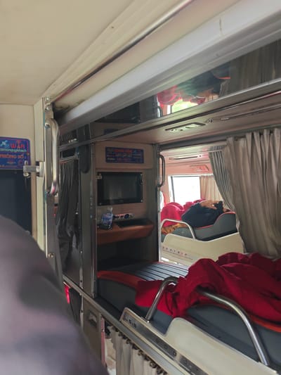 The image depicts the interior of a sleeper bus, likely in Tang Bat Ho, Vietnam, specifically showing a bunk bed section. The perspective is from a lower bunk, looking upwards and towards the right.
In the foreground, a large portion of a gray blanket or fabric is visible on the left, obscuring part of the view. The bed itself is a dark gray mattress with chrome safety rails. A bright red blanket is draped over the rails and mattress in the visible bunk.
Further back, a section of the bus's interior is visible, featuring several amenities. A small screen, possibly a television, is mounted on the wall, with a bottle of water and some buttons below it. To the left of this, a blue sign with white lettering, possibly indicating "WIFI" and other information, is attached to the wall. A metal handle is also visible on the left side of the image.
On the upper level of the bus, a person is lying down, partially covered by dark clothing. They appear to be resting or sleeping. Gray curtains are drawn on the right side, suggesting privacy for the sleeping areas. Light filters in from a window at the far end of the bus, revealing a glimpse of the outside environment, which appears to be green. The overall impression is of a compact, utilitarian sleeping arrangement within a vehicle.