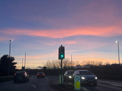 Cars travel a wet road at dusk. A traffic light glows green amidst the scene, with sparse trees silhouetted against the colorful sky. Streetlights line the roadway. The sky transitions from shades of pink and orange to blue and purple. Buildings appear faintly in the distance.