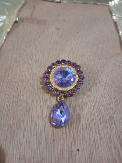 A single ornate piece of jewelry is positioned centrally on a light-colored wooden surface. The jewelry comprises a primary circular component featuring a large, faceted oval gemstone of a light purple hue, encircled by a border of smaller, faceted round purple gemstones. All stones are set in a gold-toned metal frame. A teardrop-shaped, faceted purple gemstone is suspended from the bottom of the circular component by a small gold-toned link. The underlying surface displays prominent light brown wood grain and discernible horizontal plank lines. A blurred, reflective, light-toned material is visible along the upper left edge of the frame. No individuals are present, and no actions are occurring. This scene is located in Chandragiri, Nepal.