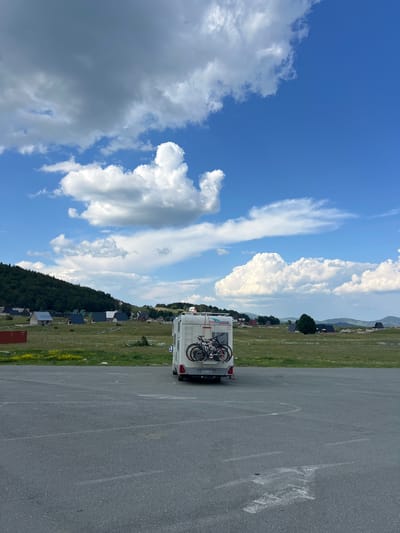The image shows a motorhome parked in an asphalt parking lot in the city of Virak, Montenegro. The motorhome is white with two bicycles mounted on the rear. In the background, there is a green field with scattered houses and a forest-covered hill. The sky is blue with scattered white and gray clouds.