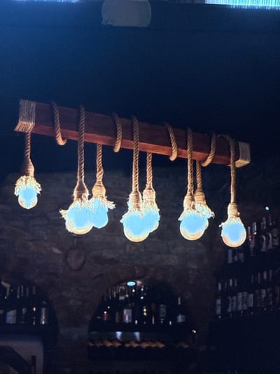 The image shows a close-up of a rustic-style light fixture with five exposed bulbs hanging from a wooden support. Each bulb is attached to the wood by a thick, braided rope that is wrapped around the wood multiple times. The ropes are frayed at the bulb connections, giving a handmade, artisanal appearance. The bulbs themselves are illuminated with a soft, blue-white light. The background is dark and appears to be a dimly lit interior space, possibly with shelves or bottles visible in the distance, suggesting a bar or a similar establishment. The overall mood is warm and inviting, created by the soft lighting and the natural materials used in the fixture. The image is a vertical shot, focusing on the details of the light fixture. There is no visible text in the image. The context of Eyüpsultan, Turkey, does not provide further visual cues within the image itself.