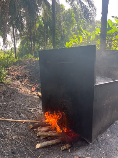 The image, rotated 90 degrees clockwise, captures an outdoor scene in Aghalokpe, Nigeria, under bright daylight. A substantial, black, rectangular metal container or vat occupies a significant portion of the frame. It is positioned horizontally across the image, with a large, bright orange and yellow fire burning intensely against its lower right side. Several pieces of wood, including logs and sticks, fuel the flames, arranged on the dark, uneven ground.

The ground is dark and appears to be a mix of soil, ash, and small debris, with some dried plant matter scattered around. A subtle wisp of steam or smoke rises from the container, particularly visible against the brighter background, indicating it is actively being heated.

Beyond the container and fire, a lush tropical environment is visible. Numerous tall palm trees, with their characteristic fronds, dominate the background, alongside other broad-leafed green plants, possibly banana trees, some illuminated by direct sunlight. The vegetation suggests a humid, fertile region. The bright light implies it is daytime, likely mid-morning or afternoon. No people or visible text are present in the image. The scene depicts a rural or agricultural heating process using an open fire and a large container.