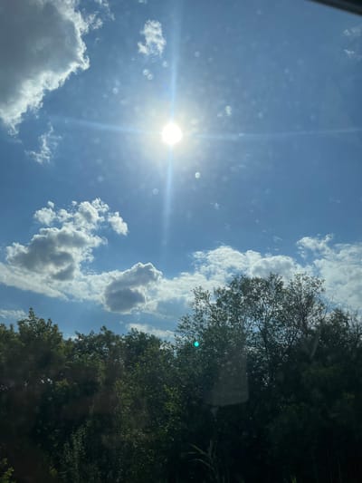 The image captures a bright, sunny day with a clear blue sky and scattered white clouds. The sun is prominently visible in the upper left quadrant, casting a strong glare and lens flare. The lower right portion of the image is dominated by the dark green foliage of trees, suggesting a wooded or park-like setting. Some of the trees appear dense with leaves, while others are more sparse, revealing the sky behind them.

The overall impression is one of a beautiful day, likely mid-morning or early afternoon given the brightness of the sun. The scattered clouds add texture and depth to the sky. There are some circular distortions visible in the sky, possibly due to reflections on a window or lens. The image appears to have been taken from inside a vehicle or through a window, given the reflections and the slight tilt of the perspective. No people, specific objects, or text are discernible in the image. The setting is natural, with a focus on the sky and trees. The weather is clearly sunny and fair.