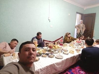 An indoor photograph captures eight individuals gathered around a long, white-tableclothed table heavily laden with food and beverages, indicative of a private residence in Samarkand, Uzbekistan.
In the immediate foreground, a male with short dark hair, wearing a grey long-sleeved shirt, holds a camera to take a selfie, looking directly at the lens. To his left, another male in a light pink polo shirt rests his head on his hand, looking down. Further back on the left side of the table, a male in a dark top is seated on a low patterned cushion, reaching towards the table. Opposite him, a woman wearing a patterned headscarf and a gold-patterned dress is seated, leaning forward. Next to her, a younger female also looks down at the table. A young child is partially visible at the far right end of the table. Another person is seated with their back to the camera on a patterned rug to the right of the table. In the background, near an open dark brown door, a woman wearing a light brown jacket and head covering stands, partially obscured, looking towards the table.
The table features numerous small plates containing various fruits (apples, grapes, bananas, pears), nuts, bread, salads, and other snacks. A large clear pitcher with red liquid, a plastic bottle of orange soda, and multiple clear glasses are also present. The room has light blue-green textured wallpaper, white crown molding, and a single light bulb hanging from the ceiling. A patterned rug in shades of red and blue covers the floor. The overall scene depicts a casual, familial meal or social gathering.
