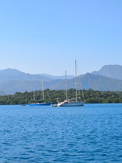 A wide, outdoor shot depicts a tranquil coastal scene in Gerokgak, Indonesia, under a clear, bright blue sky.
The upper approximately 25% of the image is occupied by a clear, pale blue sky, devoid of clouds, indicating fair weather. Below the sky, receding into the background, are multiple layers of hazy, blue-tinged mountains, exhibiting a varied, undulating topography consistent with distant landmasses. A dense, dark green treeline forms the shoreline across the mid-ground, separating the mountains from the body of water.
The lower half of the image features a calm, deep blue body of water, displaying gentle ripples and reflections of the sky, suggesting minimal wind. Two sailboats are prominently anchored or moored in the mid-ground.
The sailboat on the right, positioned closer to the viewer, has a white hull and two visible masts with rigging. The name "Pan Saga" is discernible in dark blue lettering on its port bow. A yellow inflatable dinghy is tethered near its stern. A yellow spherical mooring buoy is visible in the water slightly to its port side.
The second sailboat, located to the left and slightly further back, features a dark blue hull and at least two masts with visible rigging. A small, dark-colored tender is visible behind this vessel, closer to the treeline.
No individuals are visible on either vessel or along the shoreline. The overall environment suggests a warm, clear day at a protected bay or harbor in Gerokgak, Indonesia.