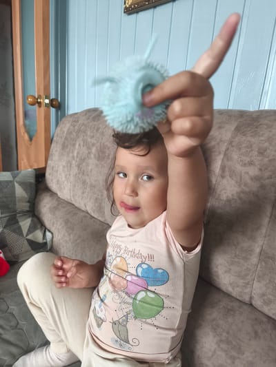A young girl is sitting on a couch, holding a blue fuzzy toy in her outstretched hand. She is wearing a pink t-shirt with colorful balloons and cupcakes printed on it, and light-colored pants. The girl's tongue is sticking out slightly as she looks to her right. The background shows a wall with vertical blue paneling and a wooden doorframe on the left. The couch is upholstered in a textured brown fabric. This image was taken in Burayevo, Russia.