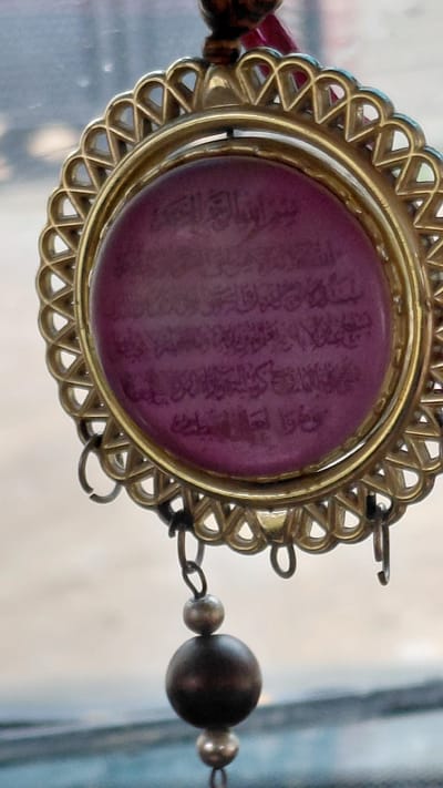 An extreme close-up shot focuses on a decorative pendant. The pendant features a circular, deep purple stone set within an ornate gold-colored metal frame. The stone is inscribed with Arabic calligraphy, which is difficult to read due to the image's resolution and the stone's texture. The pendant hangs from a thin chain and is adorned with two small, silver-colored beads and one larger, dark, spherical bead, likely made of wood or dark stone. The background is out of focus, showing a blurry suggestion of an urban or indoor environment with muted colors and light. The location is identified as Terek-Say, Kyrgyzstan, although there are no visible contextual clues within the image to support this specific geographical information.
