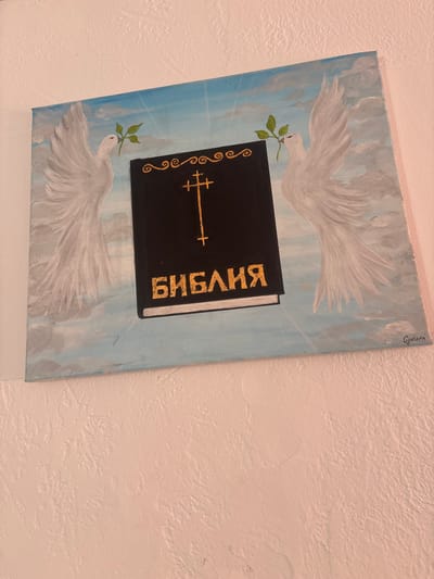 This is a painting on canvas featuring two white doves flying towards a black book. Each dove holds a green sprig with leaves in its beak. The book has the word "БИБЛИЯ" written in gold Cyrillic letters on its cover. A decorative cross symbol is also depicted on the book. The background is a sky-blue color with white clouds and streaks of light, giving the impression of a heavenly or serene setting. The painting has a roughly textured wall in the background. The artist's signature, "Гузела," is visible in the top right corner of the painting. The style of the painting appears to be religious or spiritual art.