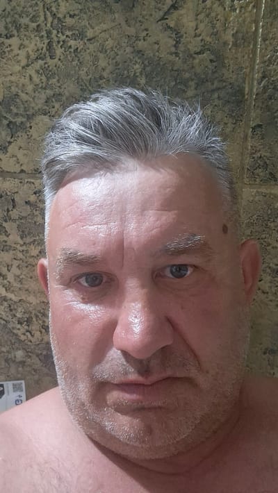 The image shows a close-up selfie of a man with graying hair and a light beard. He is looking directly at the camera with a neutral expression. The background appears to be a textured wall. There are no other people or objects of note in the image. The location information provided is not verifiable from the image itself.
