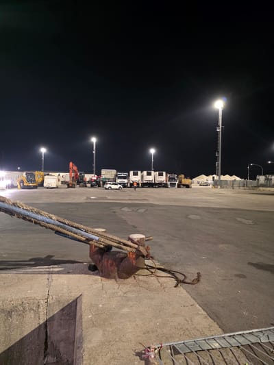 At night, in an outdoor, paved industrial area in Ravenna, Italy, several construction vehicles, including yellow excavators and an orange digger, are parked. Refrigerated trucks are lined up, and a small white car is visible. Two large industrial floodlights illuminate the scene, with one on a tall metal tower. In the foreground, thick ropes are secured to a heavy metal mooring post, attached to a concrete surface that transitions to asphalt. The overall environment appears to be a port or logistics hub.