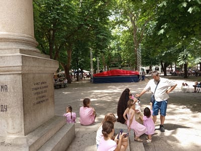 The image depicts an outdoor scene likely in Trebinje, Bosnia and Herzegovina, as indicated by the inscription on a stone monument. A large, textured stone monument is prominently featured in the foreground on the left, with some text carved into its surface. Several people are gathered in a park-like setting, under the shade of large trees.
In the middle ground, a group of children is seated and standing on stone steps leading up to the monument. Two young girls, one in a pink and white striped shirt and another in a pink dress, are sitting facing away from the camera. A third child, also in a pink outfit, is seated on the steps further to the left, facing away.
Further into the scene, a man wearing a checkered shirt and blue shorts stands and appears to be interacting with a group of children. One child, in a pink dress, stands in front of him, reaching up. Another child in a pink dress is seated to his left, also facing him. A woman with very long, dark hair is seated to the left of the man.
In the background, a large red structure is visible, possibly a stage or a display. More people are seen sitting at tables in the distance, suggesting a public gathering or outdoor seating area. The environment is characterized by abundant greenery from the trees, casting dappled shadows on the paved ground. The overall atmosphere appears to be a casual daytime event or gathering in a public space.