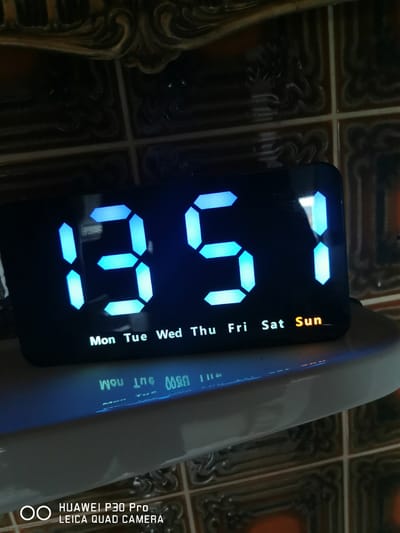 A digital alarm clock displays the time as 13:51. Below the time, the days of the week are shown, with "Sun" highlighted in orange, indicating that it is Sunday. The clock is placed on a white surface, possibly a shelf or a piece of furniture, in a room with tiled walls that have an ornate, dark pattern. The lighting in the room is dim, with the blue glow of the clock being the primary light source. The bottom of the image shows a portion of a smartphone with the text "HUAWEI P30 Pro LEICA QUAD CAMERA". There are no people visible in the image. The scene is indoors.