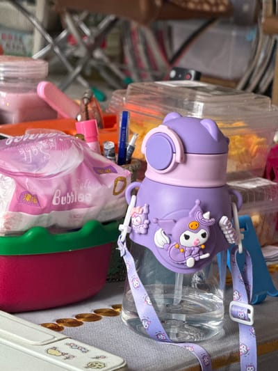 The image depicts a cluttered tabletop scene, possibly in a residential setting in Manzhouli, China. In the foreground, a purple water bottle adorned with a character resembling Kuromi from Sanrio is prominent, complete with a decorative strap. To the left of the bottle, a pink packet labeled "Soft Bubbles" is visible, suggesting it contains wet wipes or similar personal care items. Behind these items, an assortment of containers, including clear plastic ones filled with yellow food items and opaque jars, are scattered. Various small objects, such as pens and cosmetic items, are also present. The overall environment appears to be disorganized, with a mix of everyday items and decorative elements.