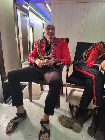 The image shows two individuals in what appears to be a dressing room.
One person, primarily visible, is sitting in a chair, wearing a red military-style jacket with gold rope details, black pants, a white head covering, and sandals. He is holding a mobile phone.
Beside him, another person is also sitting in a chair, partially visible. They are wearing a similar red jacket and black pants with a red stripe. A pair of open brown boots are on the floor by the chair.
The room seems to be a trailer/dressing room with a door and reflective panels. The flooring is light-colored. There are no elements visible in the image to indicate its location in Vamasamudram, India.