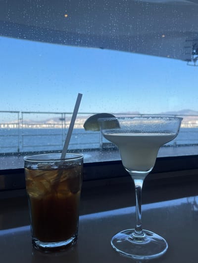 The image displays an indoor scene overlooking a maritime environment in Karşıyaka, Turkey, viewed through a window covered in water droplets. In the immediate foreground, two beverages are placed on a dark, reflective surface. The left drink is in a tall, clear glass, containing a dark amber liquid with numerous ice cubes and a white straw. The right drink is presented in a stemmed margarita glass, filled with a pale yellow liquid. Its rim is crusted with a white granular substance, and a lime wedge garnishes the rim. Beyond the droplet-covered window, a wide expanse of water, likely a gulf, stretches towards a distant, hazy shoreline or mountain range under a clear, light blue sky. A horizontal structural element, possibly a distant railing or pier, is visible over the water. The upper section of the window area shows a reflective surface, possibly an interior ceiling, with a few indistinct light reflections. No people are visible, and no actions or interactions are depicted.