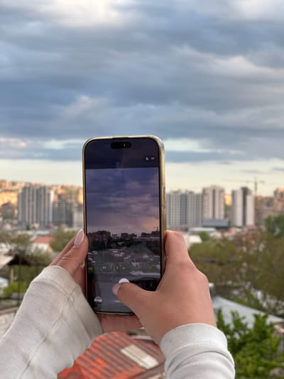 A person is holding a gold iPhone in their hands, taking a photograph of the urban landscape of Arinj, Armenia. The phone screen displays a dusk or dawn cityscape. The background beyond the phone features buildings of various heights, interspersed with trees, all under a cloudy sky. The angle of the shot suggests the person is on a higher vantage point, possibly a rooftop or elevated balcony. The person's hands are visible, with light pink nail polish. The left arm is wearing a light-colored sweatshirt with ribbed cuffs. The orange safety material is visible.