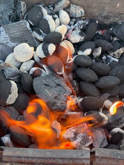 A close-up, top-down shot captures a pile of burning charcoal briquettes and larger pieces of charcoal. The briquettes are a mix of black and white, indicating they are partially lit. Flames in vibrant oranges and yellows lick across the charcoal, with a few embers glowing red. Some of the larger charcoal pieces show a cracked, ashy surface. The scene is set within a dark, metallic grill or fire pit. There are no people visible in the image. The setting appears to be outdoors, likely a backyard or park, given the natural textures of the charcoal and the implied presence of a grill. No specific location in Lunino, Russia can be identified from the image alone.