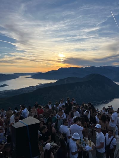 The image depicts an outdoor gathering of a large group of people at sunset, overlooking a bay with mountains in the background. The location is identified as a viewpoint near Žanjev Do, Montenegro. The sun is low on the horizon, casting a warm orange and yellow glow across the sky, which is filled with wispy clouds and contrails from airplanes.
The foreground is occupied by a crowd of people, many of whom are dressed in casual attire. They are standing and mingling, with some holding drinks and others taking photographs. Several tables are visible, laden with bottles and cups, suggesting a social event or party. A large black speaker box is positioned in the mid-ground, with blue tape around it. The people are diverse in age and gender, and their interactions appear relaxed and social.
The mid-ground shows the dramatic landscape of dark, rugged mountains sloping down to a wide expanse of water, likely a bay or fjord. The water reflects the colors of the sunset. A coastal town or settlement is visible along the edge of the water. The overall atmosphere is festive and scenic, capitalizing on the stunning natural setting and the beautiful sunset.