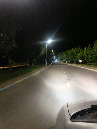 The image depicts a nighttime view from inside a vehicle driving on a road in Pivdenne, Ukraine. The road is asphalt, with white dashed lines marking the lanes and a blue and white striped curb. Streetlights illuminate the road, casting a bright glow ahead and creating a haze. Trees line the right side of the road, with a few scattered lights visible amongst them. A white car is visible in the foreground, and another vehicle is seen in the distance, its headlights on. A red and white directional sign indicating a curve to the left is posted on the right side of the road. The sky is dark and appears to be clear.