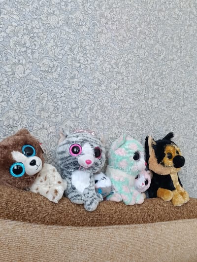 Four stuffed animals are lined up on a brown textured surface against a gray patterned wallpaper. From left to right, the animals are a brown bear with large blue glittery eyes, a grey and white striped cat with pink glittery eyes, a light green and pink spotted cat with a pink bow, and a black and tan dog with yellow glittery eyes. The stuffed animals are positioned in a row, appearing to be sitting. The background is a repeating floral pattern in shades of gray and white. The scene is indoors, with no identifiable external environment. The location is Stroitel, Russia.