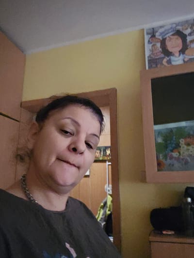 An adult woman is depicted in a close-up, sideways portrait, likely a selfie, taken indoors in Levski, Bulgaria. She is looking slightly upward and to her left with a neutral or contemplative expression. Her dark hair is pulled back, and she is wearing a dark t-shirt and a metal chain necklace.
The background shows an interior setting with walls painted in two tones: a light yellow and a lighter, possibly off-white, section. On the wall above her, two framed artworks are visible. To the left, a framed illustration features a cartoonish figure with dark, curly hair, dressed in light clothing, set against a background of other indistinct figures or elements. Further right, within a light wooden frame, is a painting or print of a vase holding a colorful bouquet of flowers, including red, blue, white, and orange blooms. To the far right, a dark electronic device and light brown cabinetry are partially visible. A wooden surface, possibly a table or desk, can be seen below her head. The lighting suggests it is daytime. No visible text is present in the image.