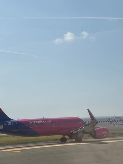 The image shows a Wizz Air Airbus A321neo airplane with registration 9H-WDQ at Fiumicino–Leonardo da Vinci International Airport near Fiumicino, Italy. The aircraft is on a taxiway or apron, likely preparing for departure or after arrival, visible because of its engine and tailfin, against a clear blue sky with contrails. The ground is paved with airport tarmac markings.