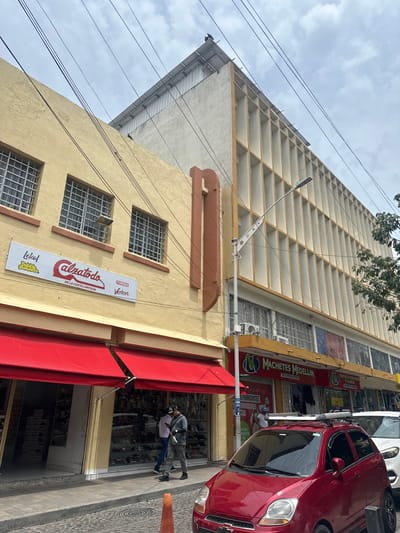 The image depicts a street scene in Perímetro Urbano Barranquilla, Colombia, featuring commercial buildings lining the street under an overcast sky. In the foreground, a red car is parked, its roof rack visible. To the left, a building with a yellow facade has a red awning over a storefront displaying shoes. A white sign above the awning reads "Lekol" and "calzatodo DE LA COSTA LIMITADA," with "avenus" and "Verlon" below. Two individuals are seen walking on the sidewalk in front of the shoe store. Further down the street, another building has a bright yellow facade with a sign for "Machetes Medellin DISTRIBUIDORA" and displays various products. The buildings have multiple windows, some with protective grates. Overhead, a network of electrical wires stretches across the frame. The street is paved with cobblestones, and a bright orange traffic cone is placed on the sidewalk.