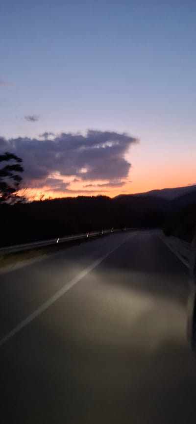 The image captures a twilight scene on a road in Valkosel, Bulgaria. The sky displays a gradient of colors, transitioning from a deep blue at the top to shades of purple, pink, and orange near the horizon, indicating either sunrise or sunset. Dark clouds are visible against the colorful sky.
The foreground is dominated by an asphalt road, with a visible white line marking the lane. The road appears to curve slightly to the right. Headlights from the perspective of the camera illuminate the road ahead, creating bright patches of light. Reflectors along the side of the road are also visible, glinting in the dim light.
A dark silhouette of trees and hills lines the horizon, separating the road from the sky. The overall atmosphere is serene and atmospheric, characteristic of dusk or dawn on a rural road. There are no people or discernible vehicles other than the implied presence of the camera vehicle.