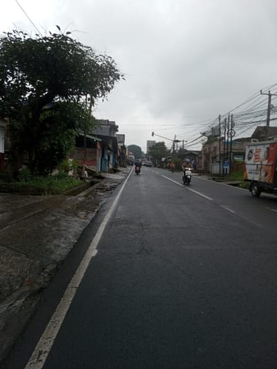 This image depicts a street scene in the city of YonPhoogla, Bhutan, under an overcast sky. The wet asphalt road, marked by a solid white line on its left edge, recedes into the distance. On the left, a large, leafy tree stands beside a concrete sidewalk and an exposed, partially damaged gutter system. Beyond these, a row of buildings, some appearing residential or commercial with varied construction materials including exposed brick, line the street.
On the road, two individuals are operating motorcycles. One rider, wearing a dark helmet and dark clothing, is on a dark motorcycle in the mid-ground, traveling in what appears to be the left lane. Closer to the viewer, another rider, wearing a light-colored helmet and light-colored attire, is on a white scooter in the right lane. Further down the street, additional vehicles are faintly visible.
On the right side of the road, a white utility truck with red and white graphics, including text and a phone number (0800 1122233), is either parked or moving slowly. Behind the truck and extending down the street, numerous utility poles with a dense network of overhead wires are present. Buildings also line the right side of the street. In the far distance, a traffic light appears illuminated green, and a large billboard structure is visible above the street level. The sky is uniformly grey with heavy cloud cover.