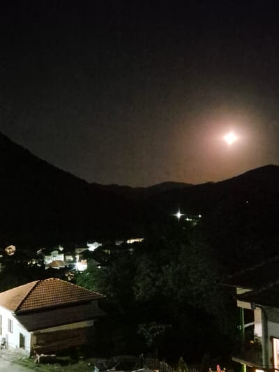 The image depicts a nighttime scene in Lesidren, Bulgaria. The dominant subject is a very bright, star-shaped light source, likely the moon, positioned high in the dark sky. Below this, a dark, silhouetted mountain or hillside rises.
Further down, the lights of a village are visible, with several houses illuminated. One house in the foreground has a distinct tiled roof, and its windows are lit, suggesting activity within. Other buildings and trees are visible in the background, also illuminated by artificial lights. There are also some streetlights visible, creating starburst effects in the image.
The scene is dark, as expected for nighttime. The bright moon and the village lights suggest a clear night, possibly with a full moon. The overall mood is quiet and serene, characteristic of a small village at night. No people are discernible in the image, and no text is visible. The perspective seems to be from a slightly elevated position, looking down upon the village.