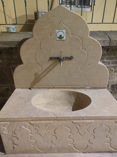 A detailed, forensic, matter-of-fact description of the image is as follows:

The image shows a marble fountain with an ornate, scalloped top. A brass faucet is mounted centrally on the upper portion of the fountain. A small, square ceramic tile with an emblem is affixed above the faucet. The emblem appears to be circular with text and imagery including a leaf. The sink basin is a smooth, carved depression in the marble countertop. The base of the fountain is a rectangular block with carved decorative elements featuring arched shapes. The fountain is situated outdoors against a stone wall. To the left of the fountain, on a stone ledge, is a white plastic container with a blue lid, likely a paint or material container. Behind the fountain, a yellow wall is visible with a green metal grate covering a window. A portion of a white ventilation unit is also visible on the yellow wall. The area appears to be a public space, possibly a park or a street in Osmangazi, Turkey, given the emblem on the tile.